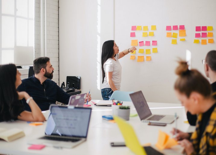 Eine Person präsentiert etwas mit PostIts an einer Wand. Vier Personen sitzen davor an einem Tisch mit Laptops.