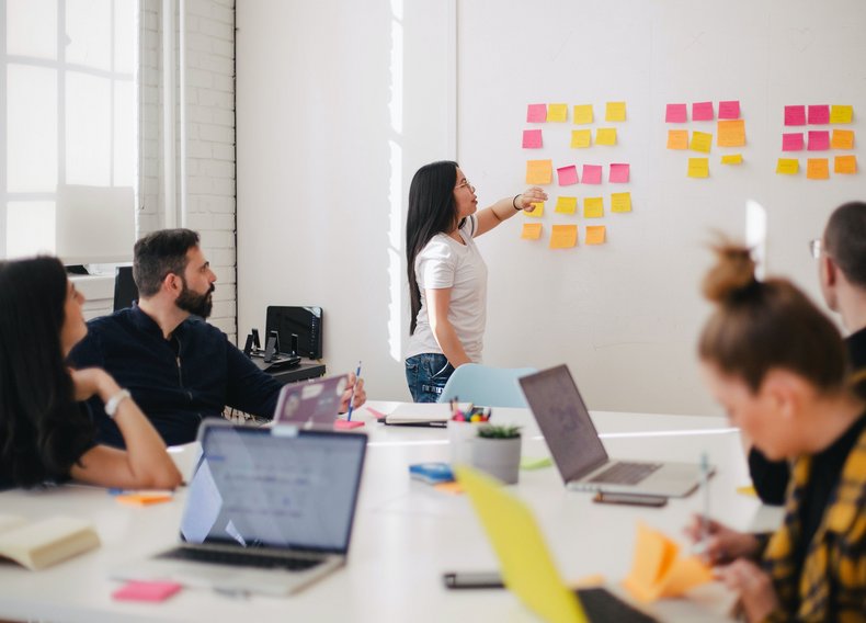 Eine Person präsentiert etwas mit PostIts an einer Wand. Vier Personen sitzen davor an einem Tisch mit Laptops.